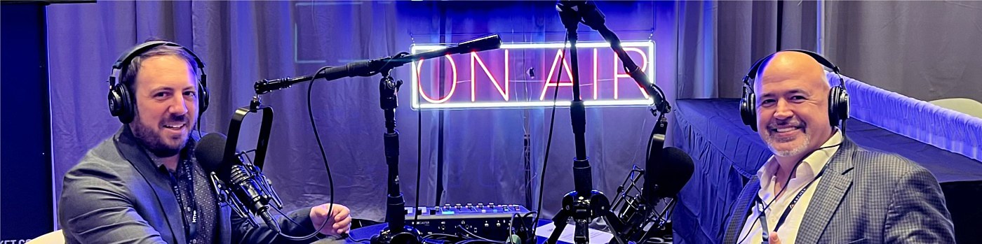 Two men seated at microphones in a podcast studio beneath a lit ON AIR sign